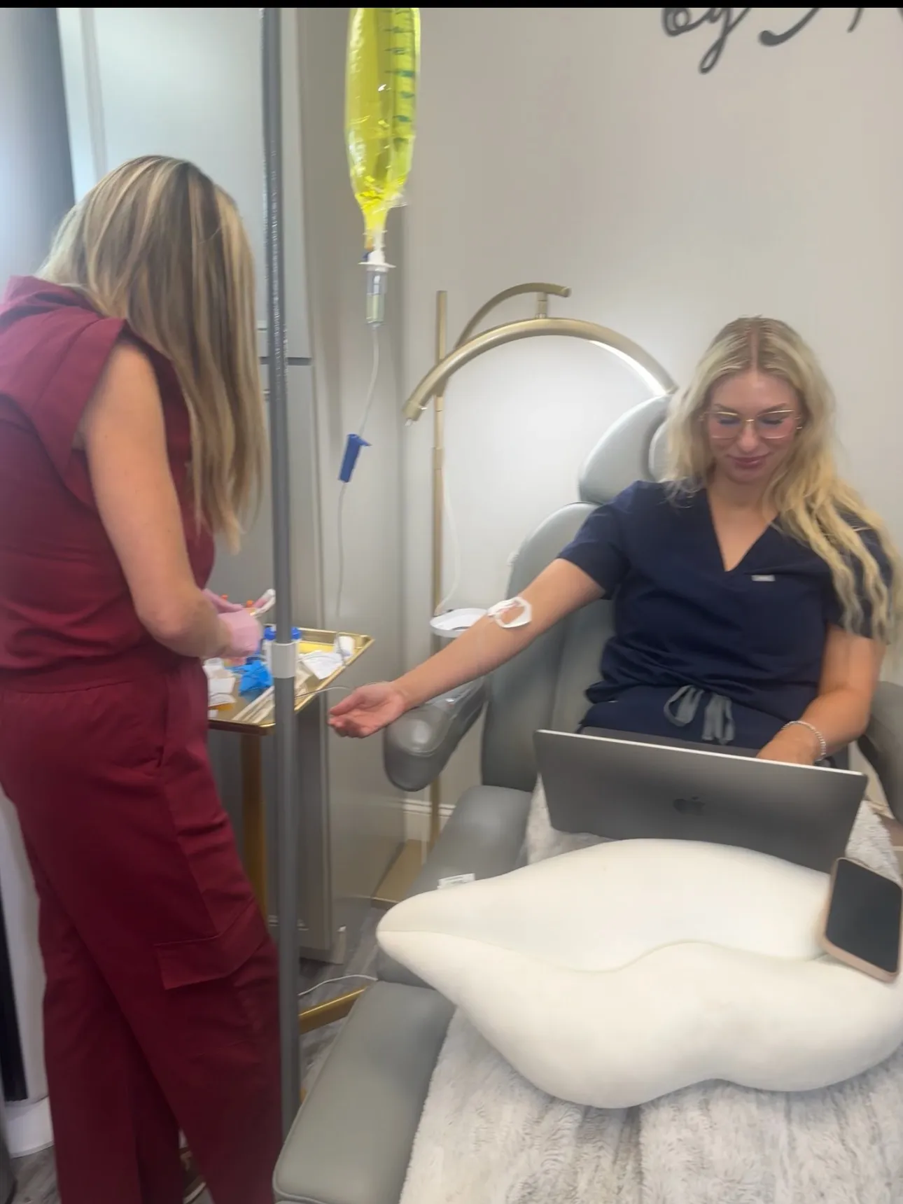 A client works on her laptop from a treatment chair during an IV infusion at Amy's Harrisburg medspa, while Amy Palacios, FNP, prepares supplies at the IV pole.
