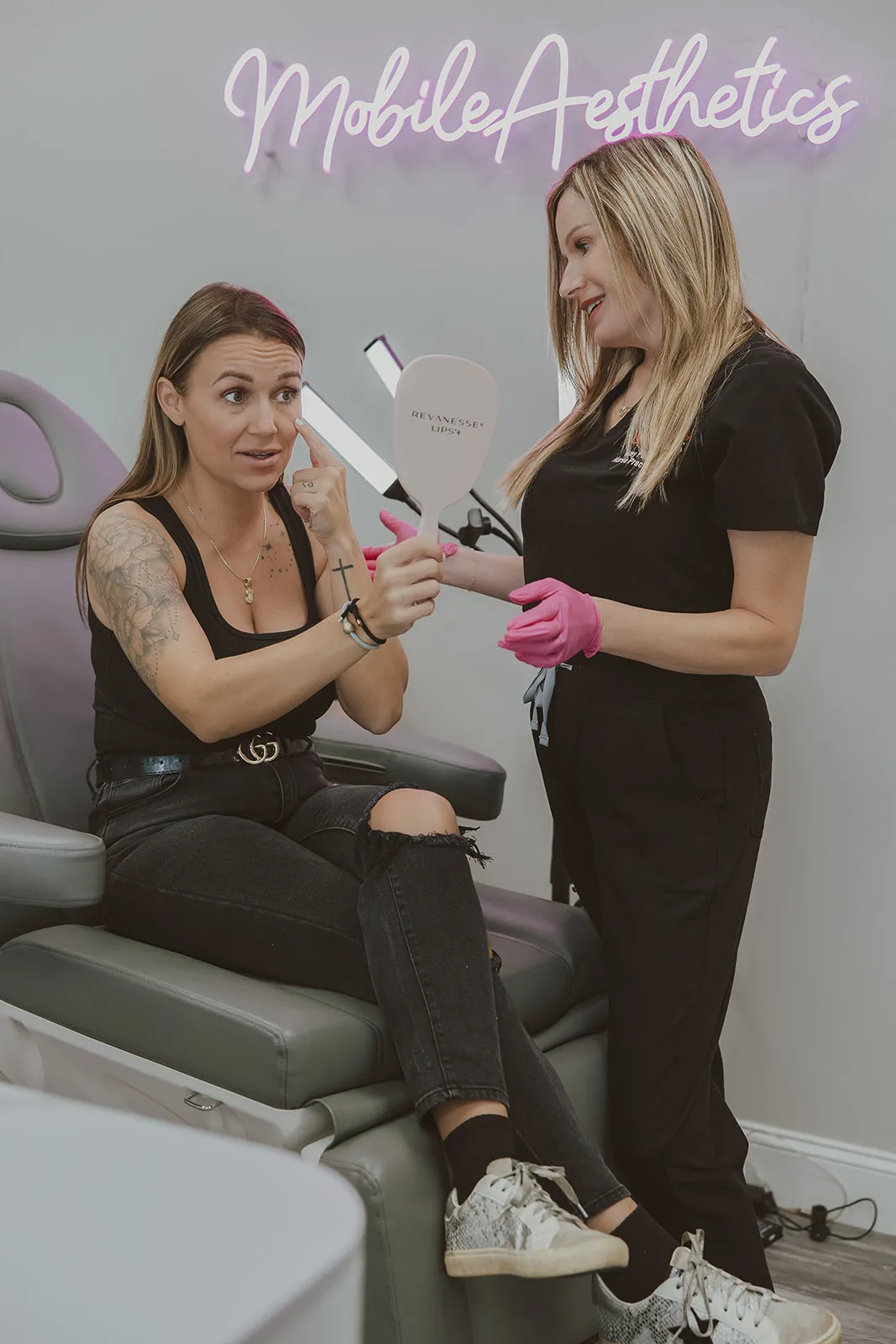 A seated client studies her face in a Revanesse Lips+ hand mirror as Amy Palacios, FNP, looks on beneath the medspa's neon sign.